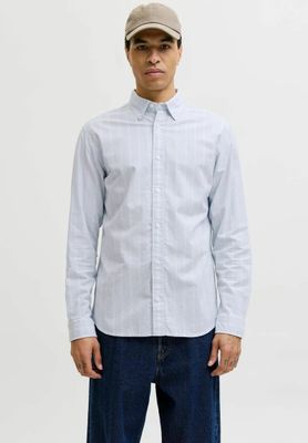 Un homme vêtu d'une chemise blanche et d'un jean bleu se tient debout devant un fond blanc.