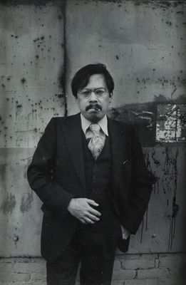 photographie en noir et blanc d'un homme en costume-cravate, debout devant un mur