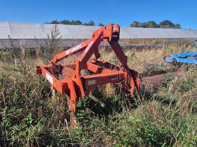 une charrue orange posée dans un champ, avec un tracteur bleu derrière elle