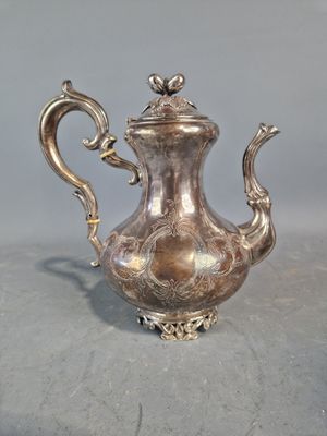 théière plaquée argent, ornée de motifs élaborés, posée sur un socle, sur fond gris