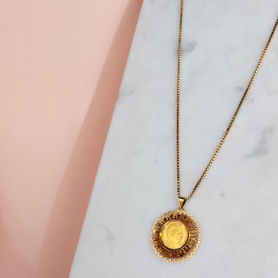 un collier avec une pièce d'or posé sur une surface en marbre, sur fond rose