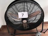 un ventilateur noir posé sur un parquet, près d'un mur blanc