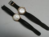 une paire de montres-bracelets noires et dorées avec des cadrans blancs, posées sur un fond blanc