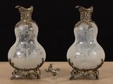 pair of white and silver vases on a table with a miniature figurine next to them