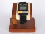 une montre numérique posée sur un socle en bois, sur fond blanc