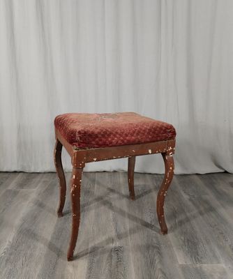 Un tabouret rembourré, dont l'assise est recouverte de tissu rouge, se trouve devant un rideau blanc.