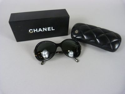 Lunettes de soleil Chanel et leur étui posés sur une surface blanche, avec une boîte noire