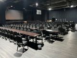 rows of chairs and tables in a large room with a projector screen in the background