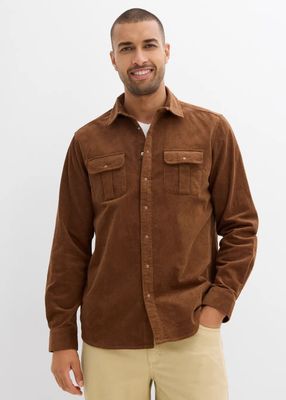 un homme debout devant un mur blanc, vêtu d'une chemise marron et d'un pantalon beige