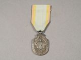 une médaille d'argent entourée d'un ruban blanc, elle-même entourée d'un ruban doré