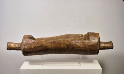 sculpture représentant une main en bois tenant un tube en bois, posée sur un socle blanc