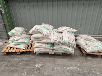 Plusieurs sacs de ciment sont posés sur des palettes devant un bâtiment