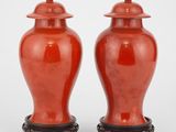 une paire de vases rouges avec couvercles, posés sur des socles, sur une surface blanche