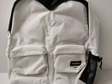 un sac à dos blanc posé sur une surface blanche, avec un sac en filet noir à côté
