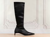 des bottes en cuir noir montant jusqu'aux genoux, posées sur un parquet devant un mur blanc