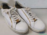 une paire de baskets blanches ornées de fermetures éclair dorées, posées sur une surface blanche
