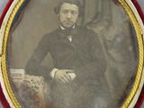 portrait d'un homme en costume noir et nœud papillon, assis sur une chaise