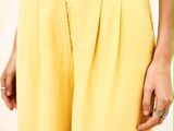 une femme vêtue d'un pantalon jaune et d'un haut argenté, debout devant un mur blanc