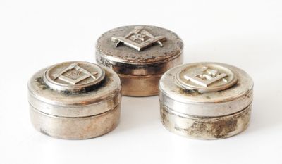 trois récipients en argent portant des initiales, posés sur une surface blanche