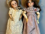 une paire de poupées anciennes, coiffées et vêtues, exposées sur un fond gris