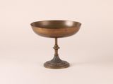 an antique brass footed bowl on a pedestal on a white background