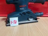 une ponceuse orbitale Bosch posée sur une table en bois, à côté d'une boîte rouge