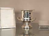 un vase en argent posé sur une table en verre, à côté d'un cadre photo