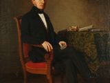 portrait d'un homme en costume noir assis dans un fauteuil rouge