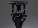 un vase noir ancien posé sur un socle, sur fond gris