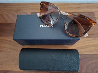 Des lunettes de soleil Prada et leur étui posés sur un parquet, devant un mur blanc