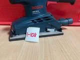 une ponceuse orbitale Bosch posée sur une table en bois, à côté d'une boîte rouge