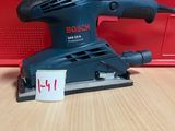 une ponceuse orbitale Bosch posée sur une table en bois, avec une boîte rouge en arrière-plan