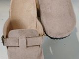 une paire de sabots en daim beige avec des boucles, posée sur une boîte en carton
