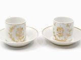 two cups and saucers with gold designs on them on a white surface
