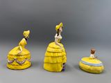 trois figurines représentant des femmes en robe jaune, assises sur une boîte jaune