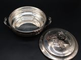 two silverplated dishes on a black surface, one with a lid and the other with a flower decoration