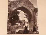 an old black and white photo of a man sitting on a bench in front of a stone arch