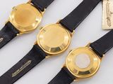 three gold watches with black leather straps and tags on a white surface