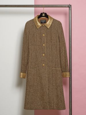 un manteau en tweed marron, avec un col et des poignets dorés, suspendu à un cintre devant un mur rose
