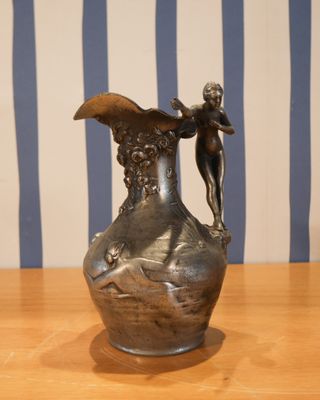 un vase en argent posé sur une table en bois, avec en arrière-plan un mur rayé de bleu et de blanc