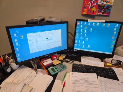 deux écrans d'ordinateur posés sur un bureau, avec des papiers et une calculatrice