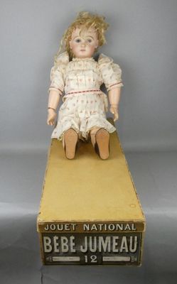une vieille poupée assise sur une boîte portant le nom de « National Bébé Jumeal »