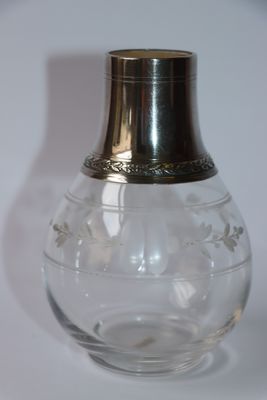 un vase en verre muni d'un couvercle en argent, posé sur une surface blanche
