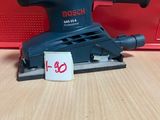 une ponceuse orbitale Bosch posée sur une table en bois, à côté d'une boîte rouge