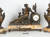 une statue représentant une femme assise sur une table, à côté de trois vases et d'une horloge
