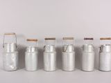 six metal milk cans with wooden handles lined up against a white wall
