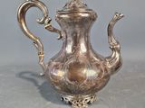 théière plaquée argent, ornée de motifs élaborés, posée sur un socle, sur fond gris