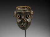 une image représentant un masque en bronze posé sur un socle, sur un fond blanc