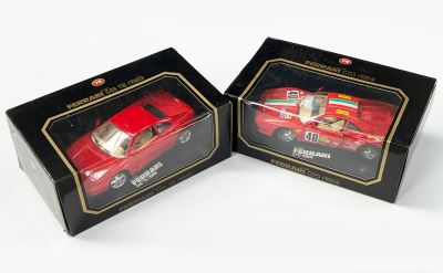 une paire de petites voitures rouges moulées sous pression, dans des boîtes noires, posées sur un fond blanc