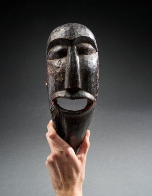 une personne tenant un masque en bois à la main, sur fond gris
